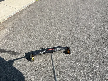 First-person view of a black magnetic sweeper with yellow wheels and a red-gripped handle resting on an asphalt driveway near a white Ford truck and utility trailer