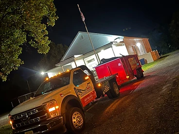 White Ford Super Duty truck hitched to a red debris trailer parked on a gravel driveway at night with an American flag flying on a flagpole behind