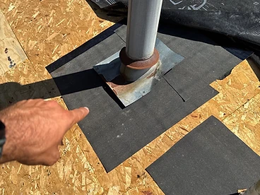 Hand pointing at a square patch of black ice and water shield membrane around a metal plumbing vent pipe boot on a plywood roof deck