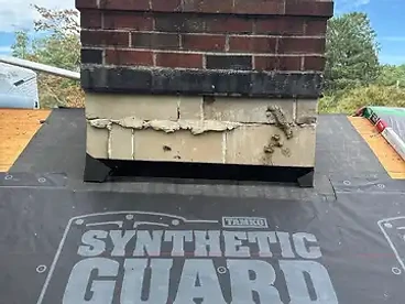 Black TAMKO synthetic underlayment installed up to the base of a brick chimney on a roof deck with exposed OSB on either side