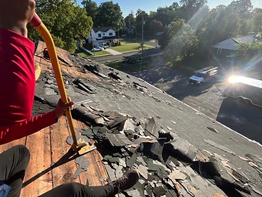 First-person view down a roof slope showing a worker prying up old shingles to reveal the weathered wooden sheathing below