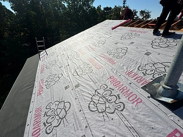 High-angle view of a roof covered in white Owens Corning ProArmor underlayment with pink branding overlapping toward the eave with a metal chimney pipe boot