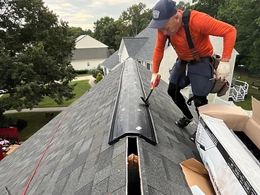 Roofer in an orange shirt and blue cap holding a hammer to secure a black ridge vent to the peak of a grey shingled roof with a vent strip box nearby