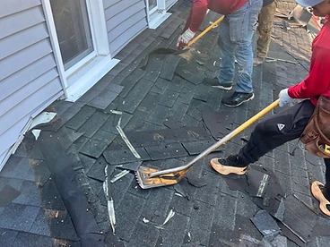 Three workers in red shirts removing weathered asphalt shingles with roof shovels against a grey vinyl sided wall