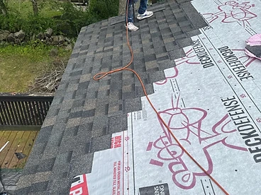 Roofer in a red shirt standing on installed architectural shingles holding a nail gun with Deck Defense underlayment still exposed above and an orange air hose trailing across the roof