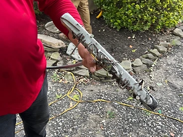 Roofer in a red shirt holding out a silver-handled magnetic sweeper head coated in collected nails and metal debris beside an evergreen bush