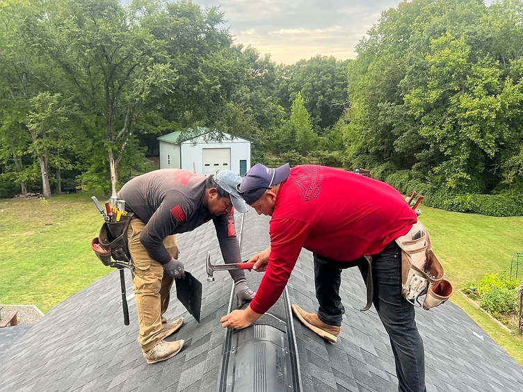 Two roofers in red Veterans Roofing shirts installing black ridge cap shingles over a ridge vent on the peak of a charcoal shingled roof