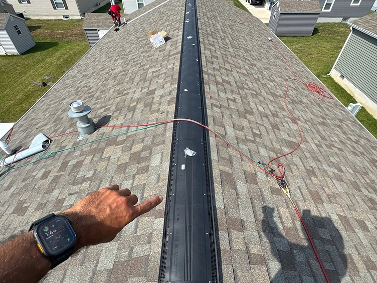 First-person view down a roof ridge line with a black ridge vent installed along the peak and a hand pointing toward it