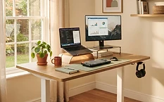 A warm, sunlit home office with a modern electric standing desk in mid-standing position, natural wood surface, laptop and monitor, terracotta and sage accents
