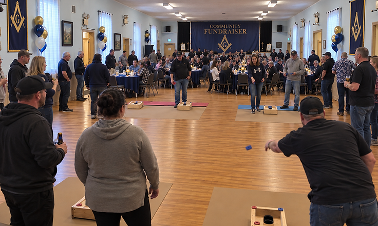 Cornhole community fundraiser in the lodge hall, with a Masonic square-and-compass banner reading Community Fundraiser and guests gathered at navy-clothed tables