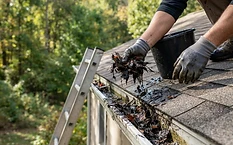 Gloved hands scooping wet leaves out of a white residential gutter