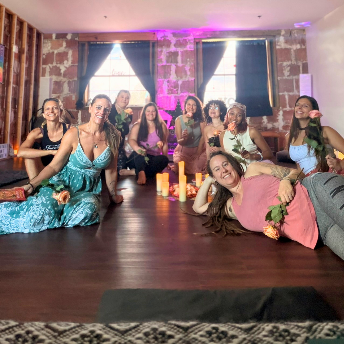 Ten women relaxing together after a somatic class on the studio floor, most holding pale orange roses, lit by candles and a soft purple glow