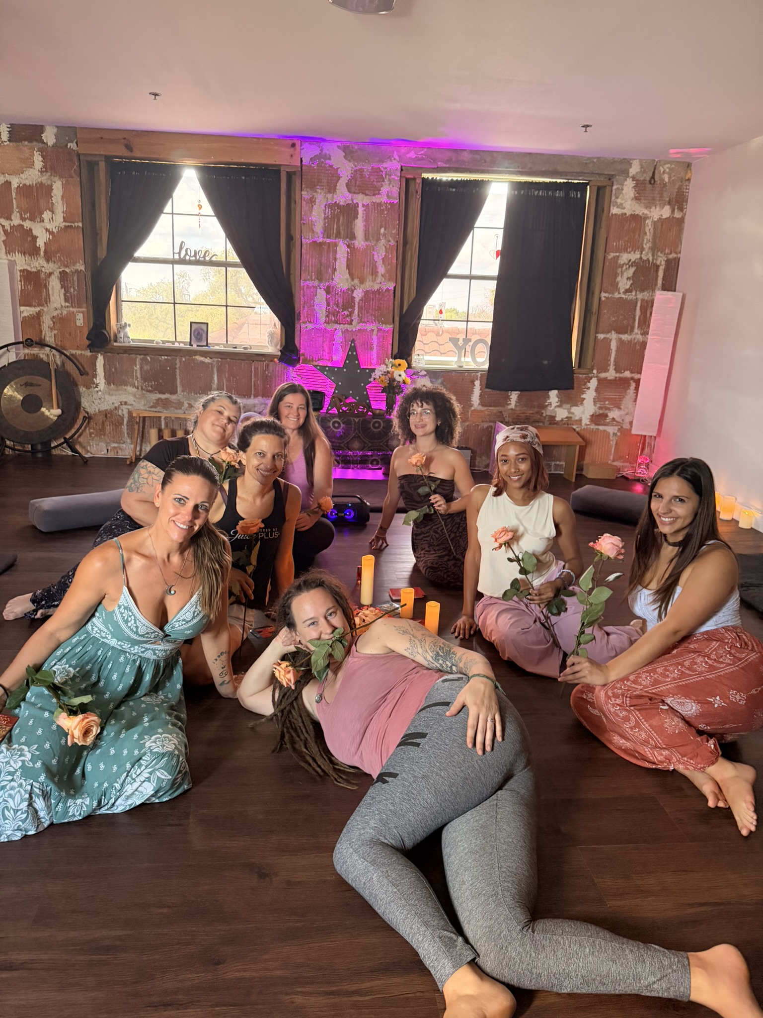 A relaxed circle of women on the studio floor holding roses, lit by candles and a soft purple glow