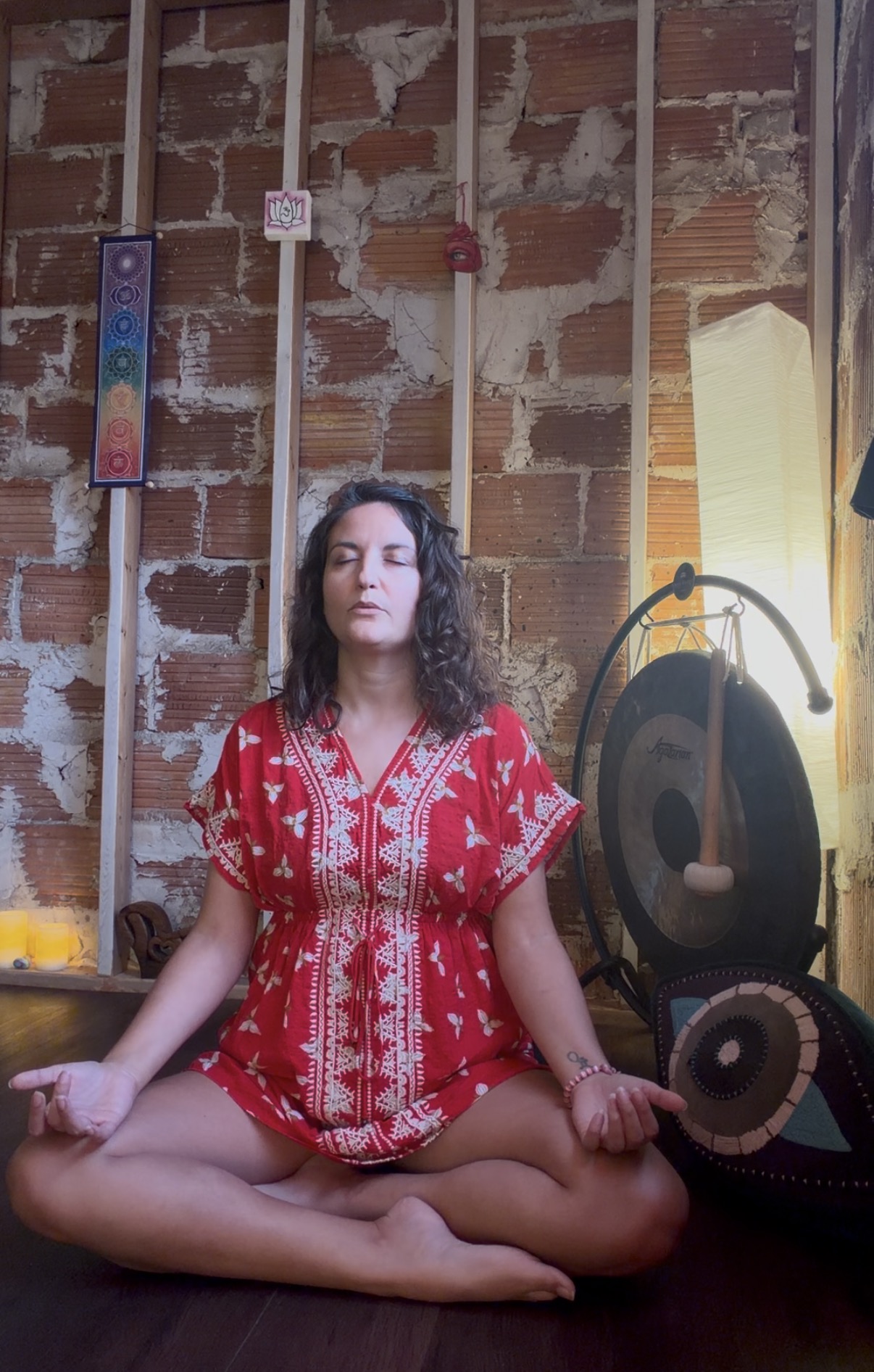 Brittany meditating cross-legged beside a large gong, wearing an embroidered red dress in the candlelit studio