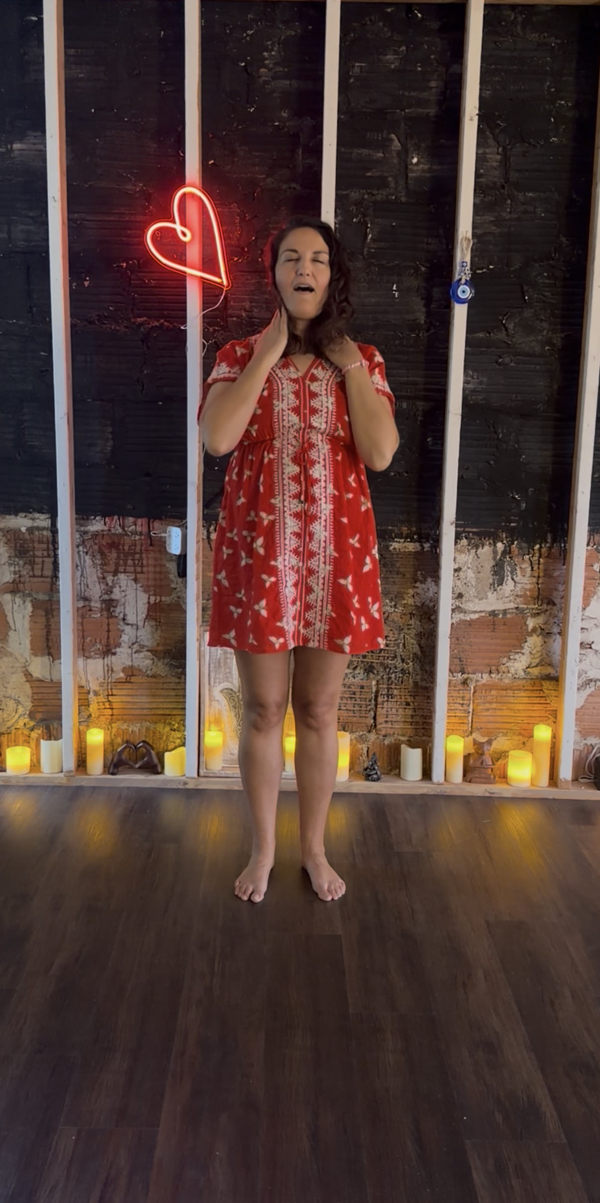 Brittany standing barefoot with her eyes closed and hands resting near her throat, demonstrating somatic movement in front of a glowing neon heart