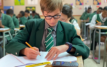 A South African high school learner in school uniform writing in a notebook at a desk with textbooks, stationery and a calculator
