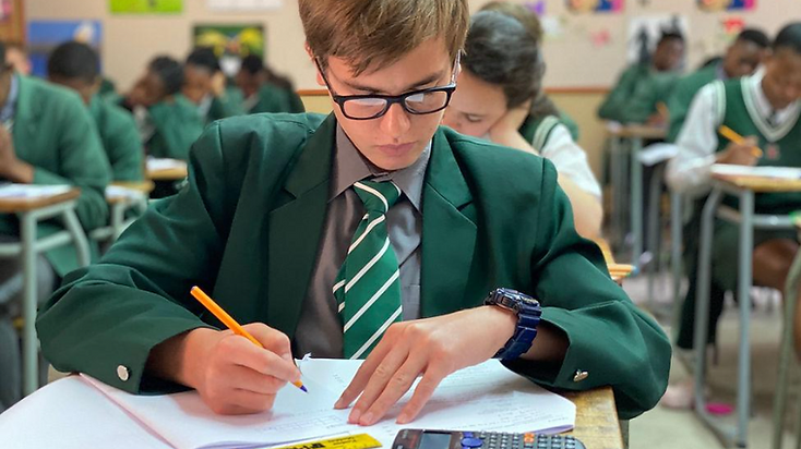 A South African high school learner in school uniform writing in a notebook at a desk with textbooks, stationery and a calculator