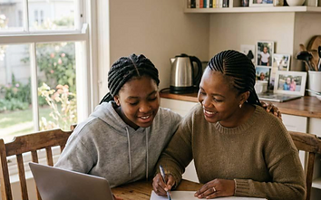 A South African parent sitting with her daughter at a kitchen table, helping her with homework while she works on a laptop