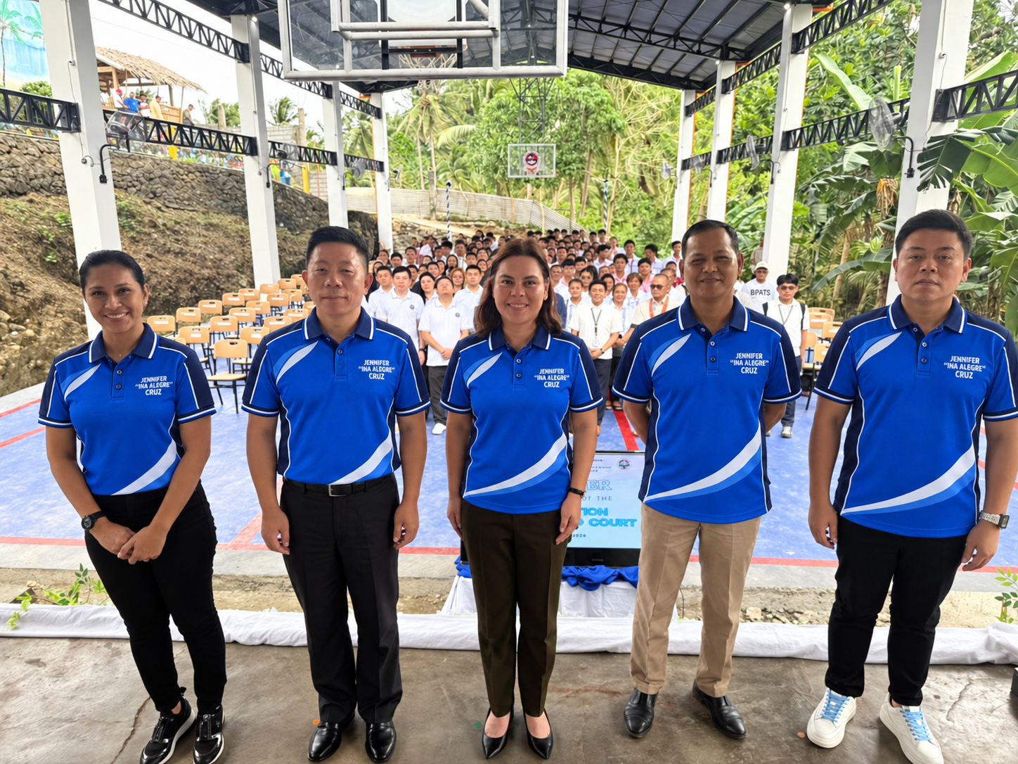 Five officials in blue shirts standing side by side on a newly finished covered court, with a seated audience behind them.