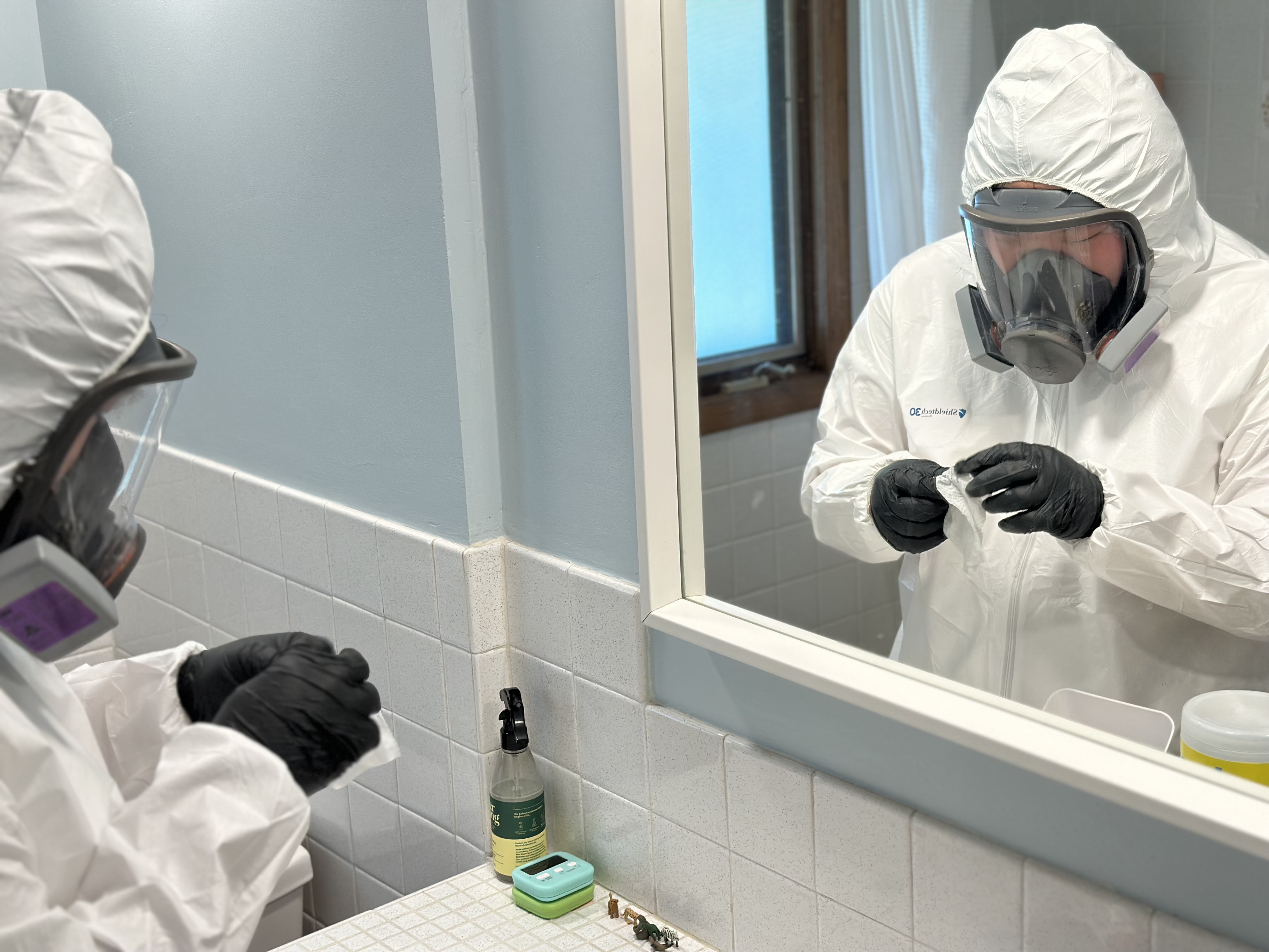 Abatement technician cleaning near a bathroom mirror in full respirator