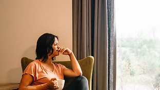 A woman sitting thoughtfully in an armchair by the window, holding a mug