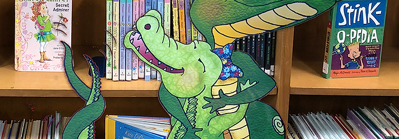 KidLit Librarian mascot alligator among library bookshelves