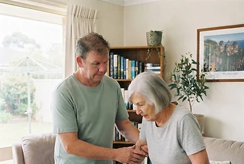 Warren, a support worker, patiently helping an older participant with a walking frame in a bright living room