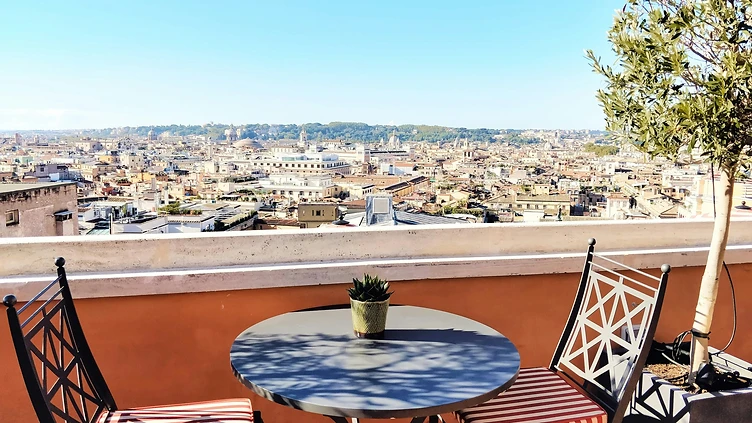 Terrazza panoramica di un attico a Roma con tavolino, sedie in ferro battuto e ulivo, vista sui tetti e sulle cupole del centro storico