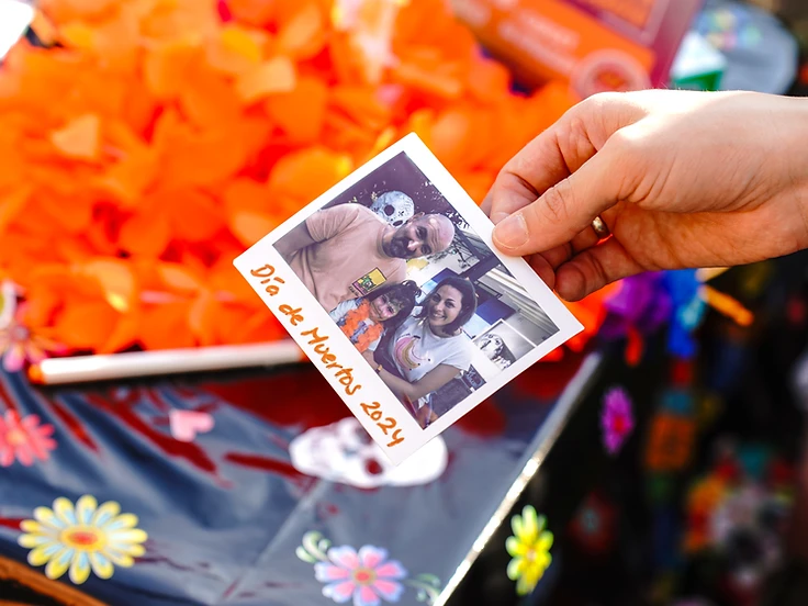 Una mano regge una fotografia istantanea di una famiglia sorridente tra i fiori di cempasúchil, durante il Día de los Muertos Roma.