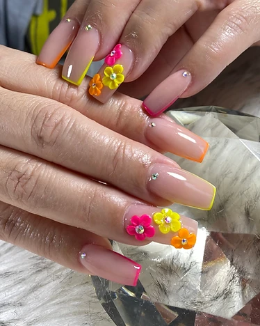 Long square nails with nude base and orange, yellow, and pink French tips with 3D flowers