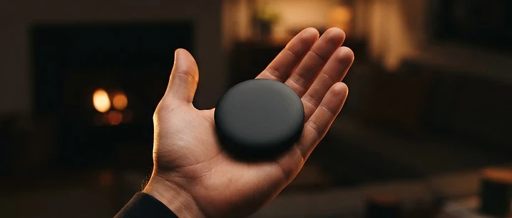 Small black streaming puck held in an open palm against a blurred dark living room