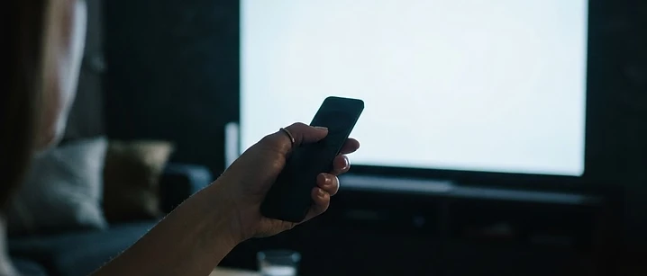 Hand holding a remote pointed at a large blank glowing television screen in a dark room