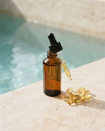 An unlabeled amber glass bottle of golden fish oil beside translucent golden softgels on a cream stone surface.