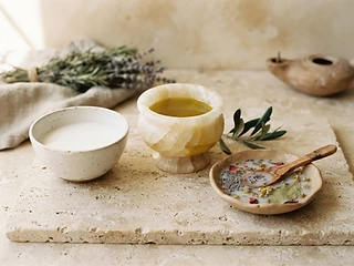Still life of olive oil in an alabaster vessel, a bowl of milk, rice water, and dried herbs on travertine