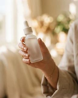 A hand holding an unlabeled frosted glass skincare bottle in soft natural light