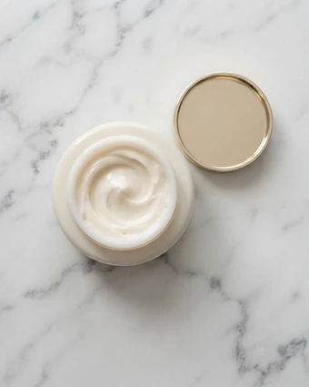 Open white cream jar with a smooth swirl of ivory moisturizer and a champagne lid on marble