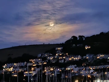 A full moon rising over the lit hillside and moored yachts on the Dart