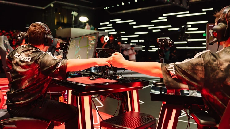 Two players fist-bump across their desks during a match on an esports stage.