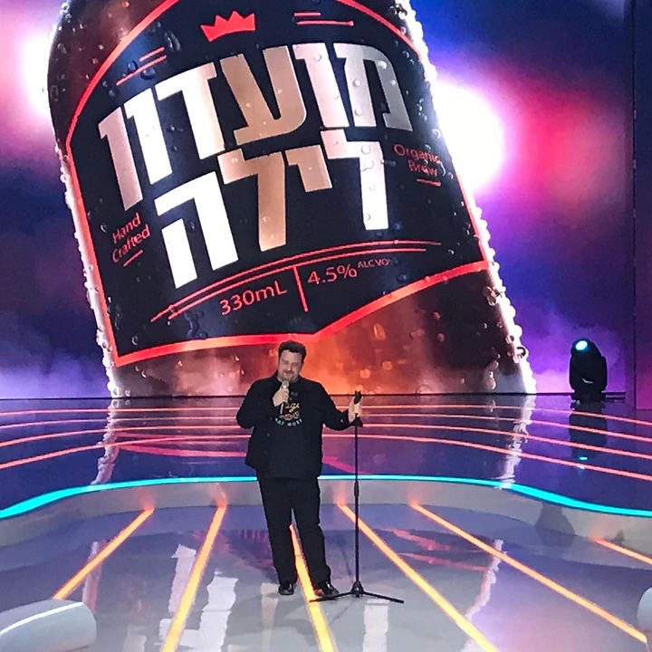גידי ליבנה מופיע כסטנדאפיסט על במה עם רקע גדול של מועדון לילה.