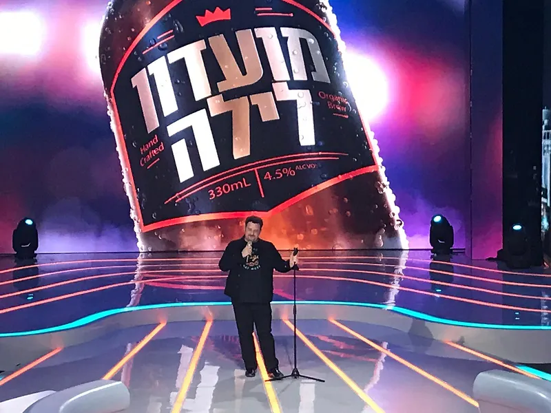 גידי ליבנה מופיע כסטנדאפיסט על במה עם רקע גדול של מועדון לילה.