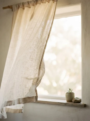 A sheer cream linen curtain billowing in a breeze in front of a sunlit window