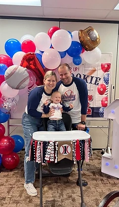 A family celebrating a baby's first birthday with a baseball-themed balloon arch