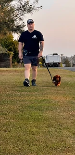 Trainer walking a dachshund on lead across a grassy field at golden hour