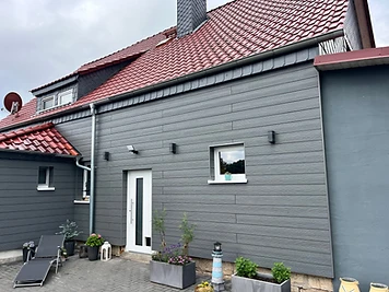Haus mit grauer Holzfassade, weißer Haustür und rotem Ziegeldach