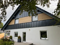Zweigeschossiges Haus mit Holzlamellen-Fassade, anthrazitfarbenen Paneelen und überdachtem Sitzplatz