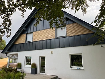 Zweigeschossiges Haus mit Holzlamellen-Fassade, anthrazitfarbenen Paneelen und überdachtem Sitzplatz