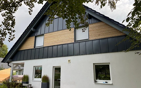 Zweigeschossiges Haus mit Holzlamellen-Fassade, anthrazitfarbenen Paneelen und überdachtem Sitzplatz