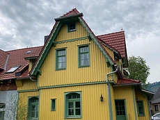 Zweigeschossiges Wohnhaus mit gelber Holzfassade, grünen Fensterrahmen und rotem Ziegeldach