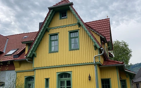 Zweigeschossiges Wohnhaus mit gelber Holzfassade, grünen Fensterrahmen und rotem Ziegeldach