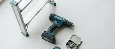 Flat lay of a cordless drill, step ladder and measuring tape on a bright surface